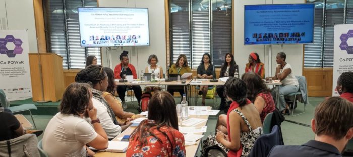 A photo taken at the Launch event. It shows event guests seated in the foreground looking at the panel of speakers sat at a table in front of two screens. The panel consists of eight women of colour - seven professors from the Co-POWeR team and host, Naz Shah, Labour MP for Bradford West.